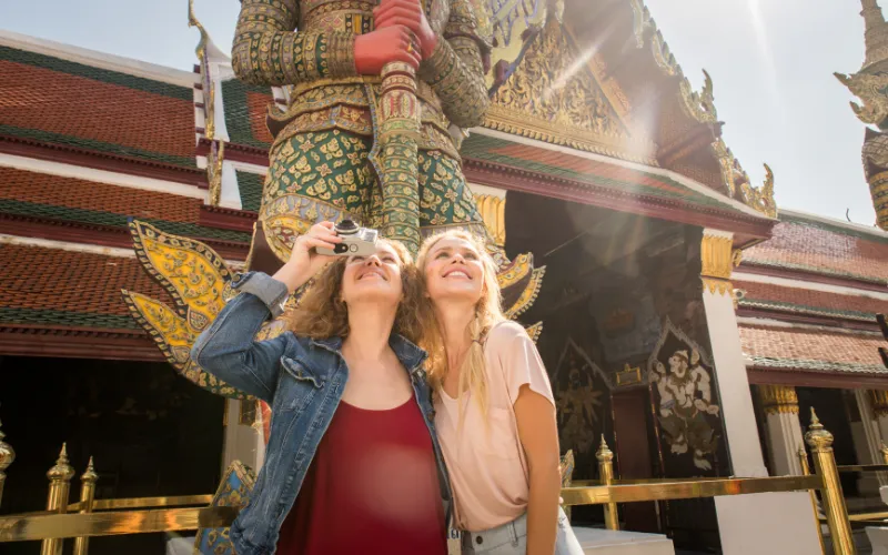 Two smiling tourists exploring the Grand Palace in Bangkok, Thailand. They are admiring the intricate details of a giant Yaksha guardian statue while capturing memories with a camera under the bright sunlight.