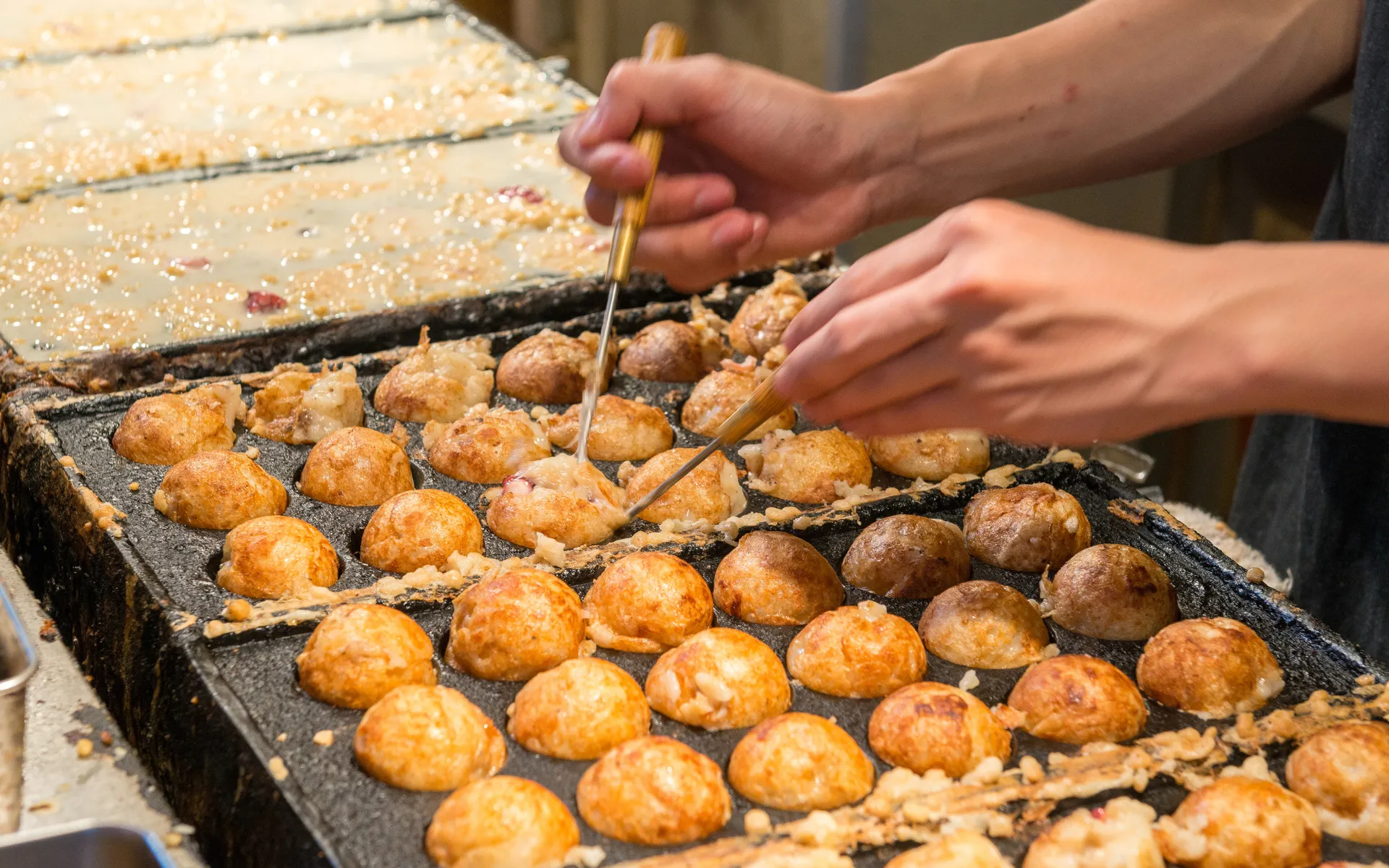 Hands preparing traditional Japanese takoyaki octopus balls on a hot griddle.