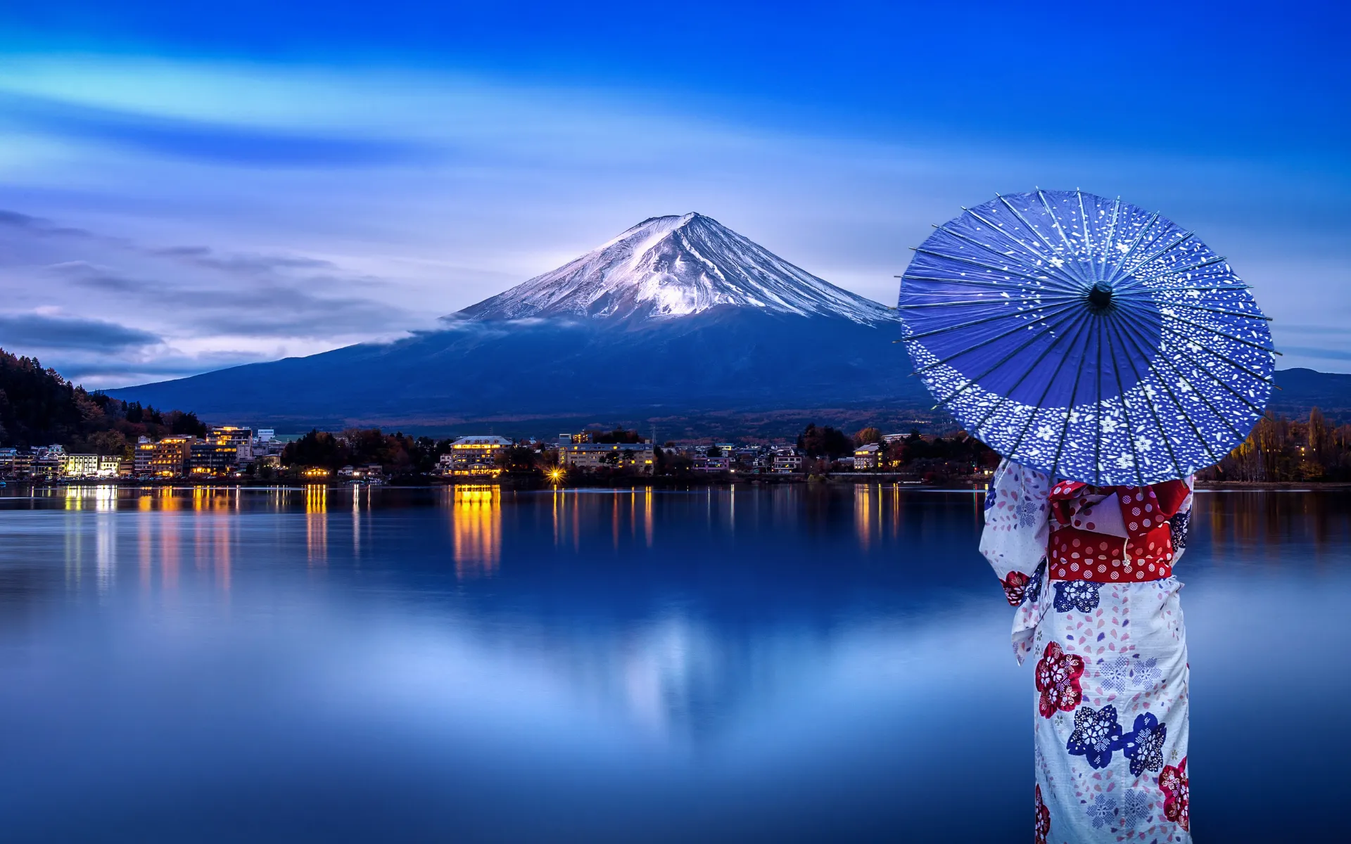 Woman in a traditional kimono holding a Japanese parasol gazes at snow-capped Mount Fuji across a serene lake at dusk, with city lights reflecting on the water.