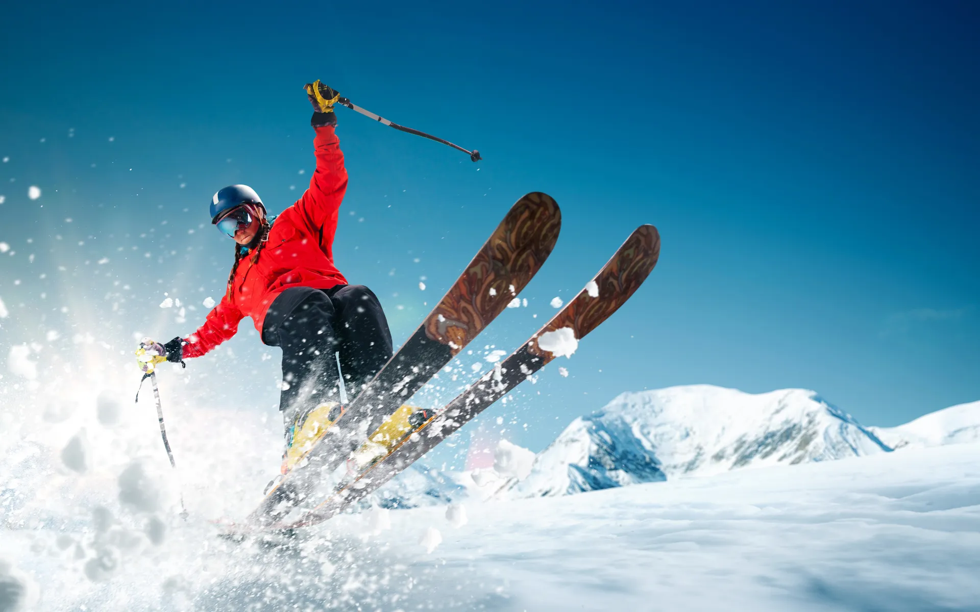 Skier in red jacket making a dynamic turn on a snowy slope with snow flying, set against clear blue skies and snow-covered mountains in the background.