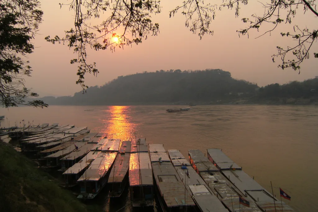 Golden sunset reflecting on a serene river in Laos with traditional longtail boats moored, evoking the tranquil beauty of luxury river cruises and exclusive event settings.