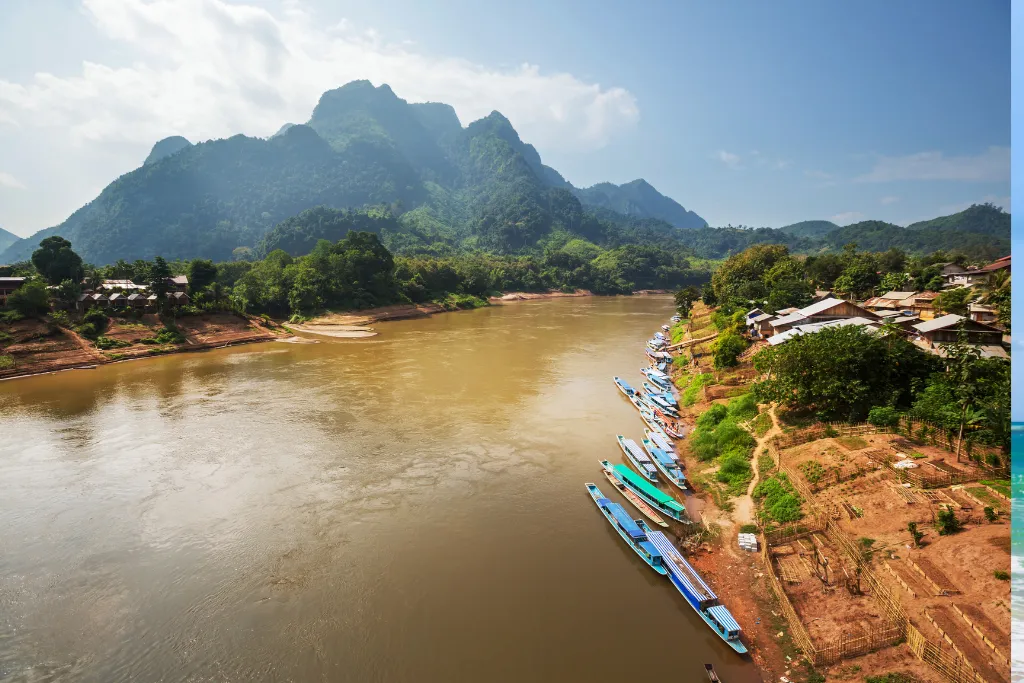 Panoramic view of a wide river with traditional boats and a riverside village in Laos, highlighting cultural immersion and scenic tours for luxury groups.