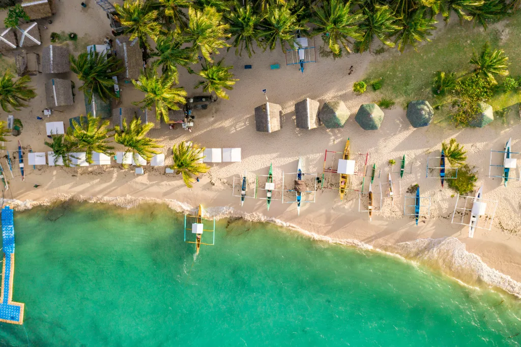 Top-down view of a tropical beach in Siargao with palm trees, bungalows, and traditional boats, highlighting a luxurious and secluded retreat for MICE groups and individual travelers in the Philippines.