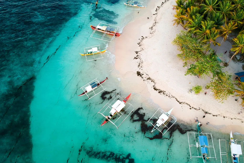 Overhead view of a pristine white sand beach in Siargao with traditional banca boats on turquoise waters, showcasing a luxury resort destination for discerning travelers and MICE groups in the Philippines.