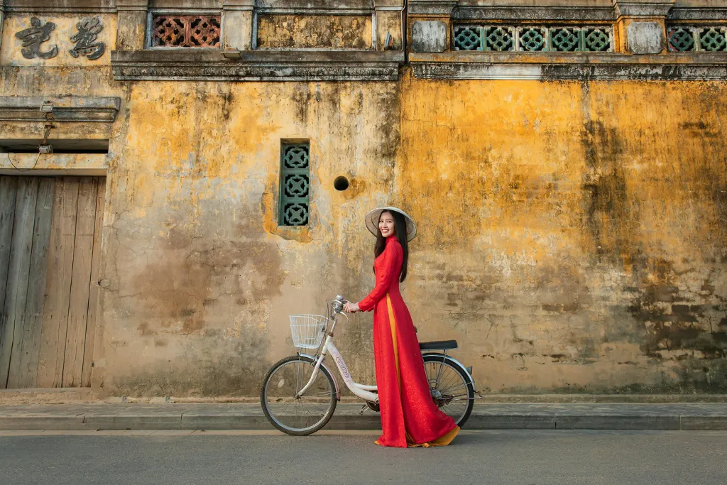 A graceful woman in a traditional Vietnamese Ao Dai with a bicycle against an ancient yellow wall in Hoi An, symbolizing immersive cultural experiences for luxury travelers.
