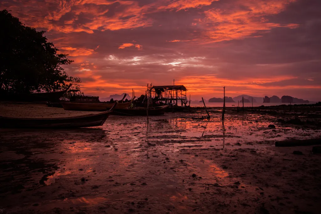 A dramatic and exotic sunset reflecting on the water, with traditional Thai boats and a pier, representing a unique and unforgettable luxury travel experience.
