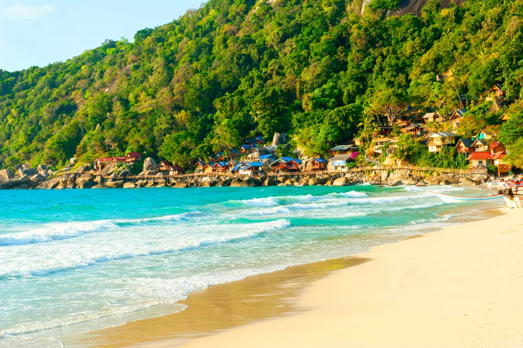 A view of a beautiful white sand beach with turquoise water and a line of colorful beachfront bungalows nestled against a lush green hillside in Ko Pha-ngan, Thailand. This image captures a perfect luxury island escape.