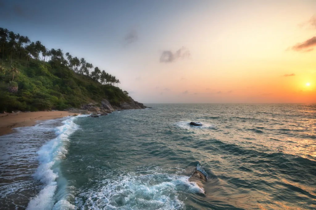 A breathtaking sunset over the ocean on a secluded beach in Ko Pha-ngan, Thailand. The view features a lush hillside with palm trees, gentle waves, and a beautiful golden sky, perfect for a luxury escape.
