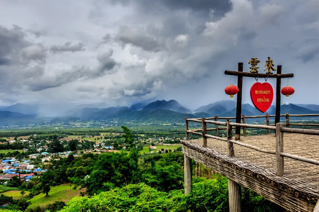 The Yun Lai Viewpoint in Pai, with a wooden deck and a prominent heart-shaped sign, overlooks a sprawling valley and distant mountains under a dramatic sky. This iconic spot is a must-visit for luxury tourists and a highlight for any DMC.