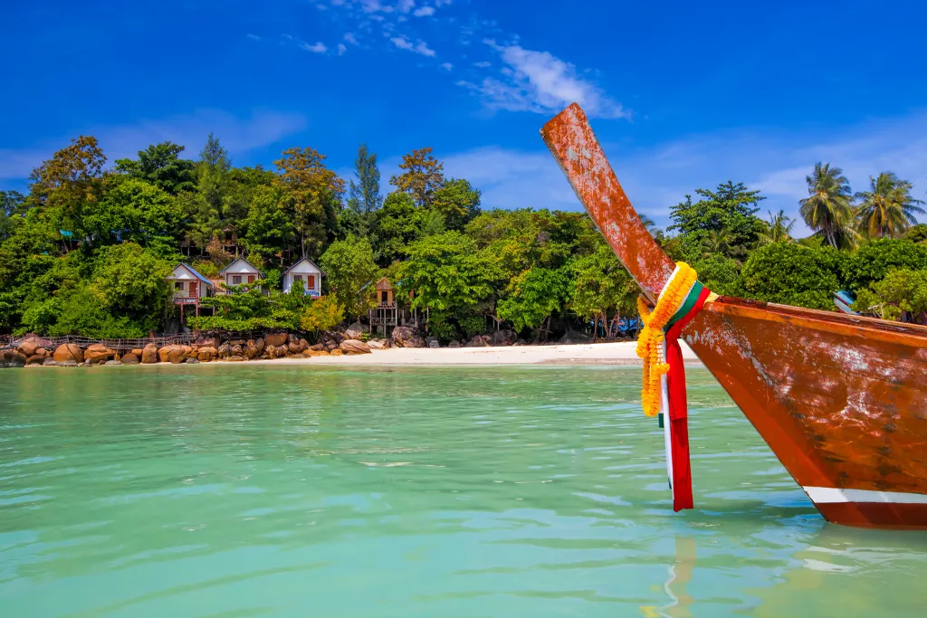 The ornate bow of a traditional longtail boat on the calm, clear turquoise waters of Koh Lipe, Thailand, with a stunning beachfront resort nestled in lush greenery in the background. This image represents a luxury beach vacation and private boat excursion.