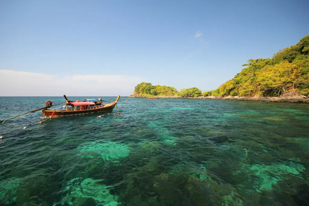 A traditional wooden longtail boat with a red roof floats on turquoise, crystal-clear waters near a lush green island in Koh Lipe, Thailand. The image represents a luxury beach vacation and private excursion.