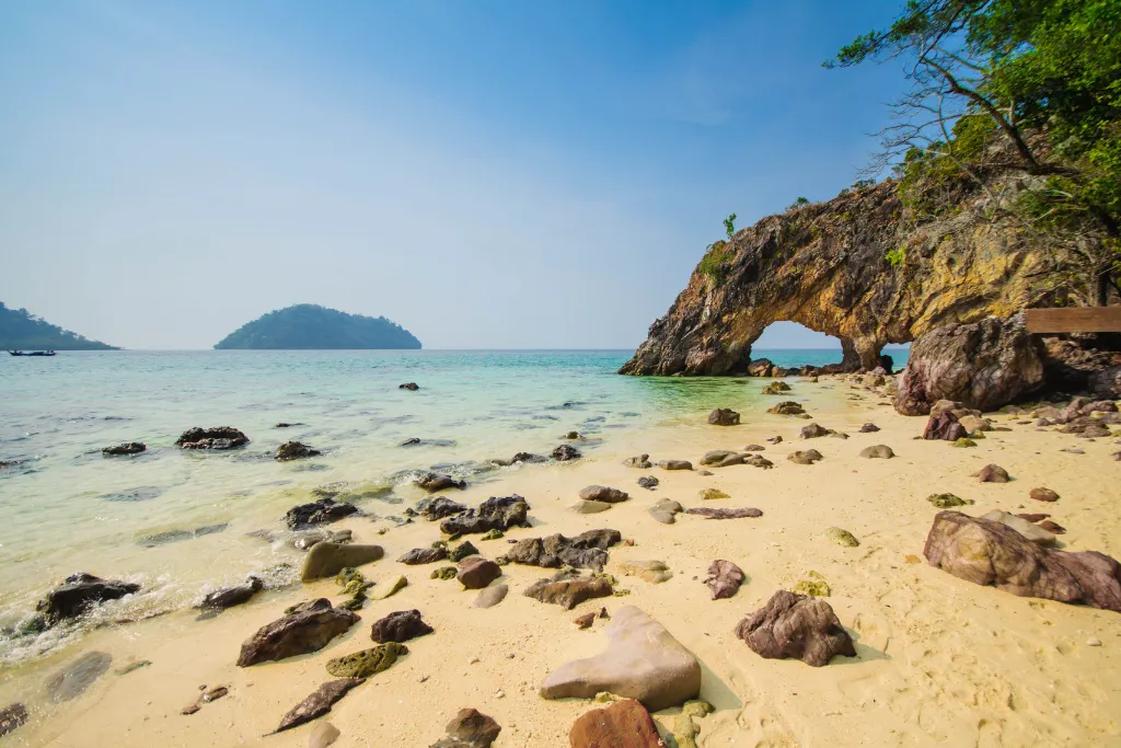 A scenic view of a unique rock arch on a tranquil beach in Koh Lipe, Thailand. The sandy shore is scattered with rocks, and the calm turquoise water meets the lush green coastline, highlighting a unique and luxurious travel destination.
