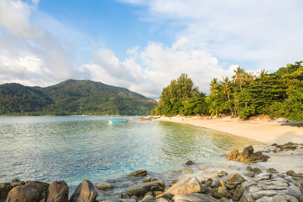 A stunning tranquil bay in Koh Lipe, Thailand, with clear, calm water and a sandy beach lined with lush green palm trees and tropical foliage. A distant mountain range and a small boat add to the peaceful, idyllic scene, perfect for a luxury travel escape.