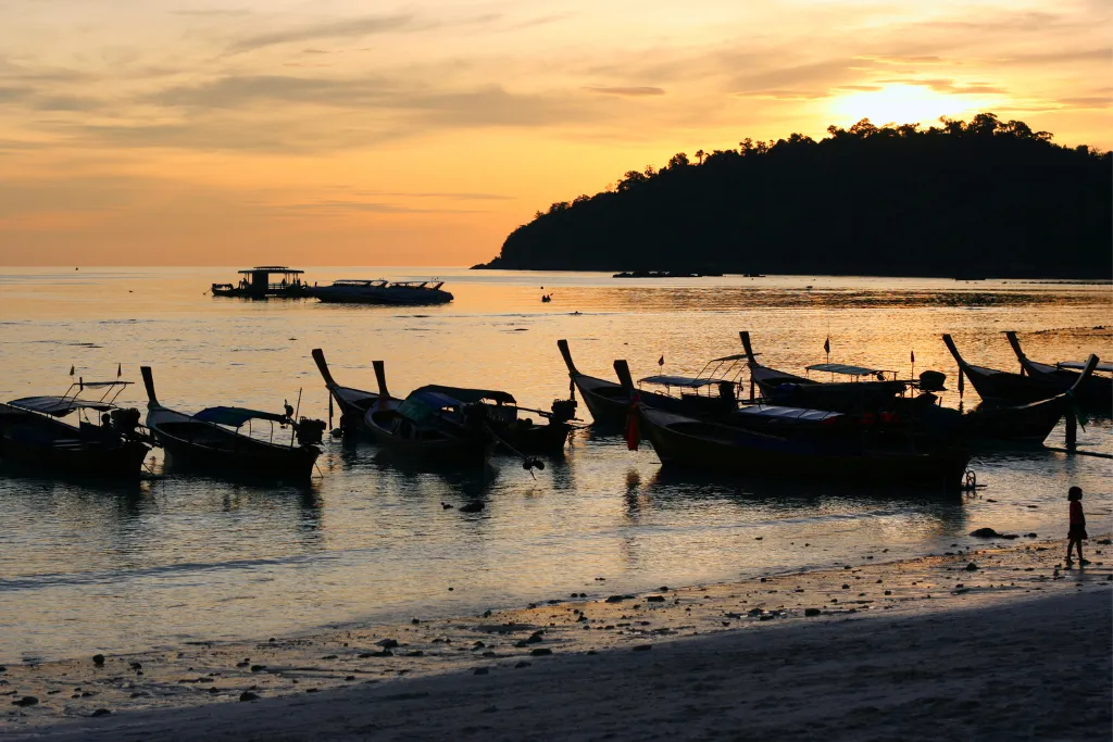 A stunning sunset silhouettes traditional Thai longtail boats anchored on the beach in Koh Lipe, Thailand. The warm orange and yellow sky reflects on the calm water, creating a serene atmosphere perfect for a luxury travel experience.