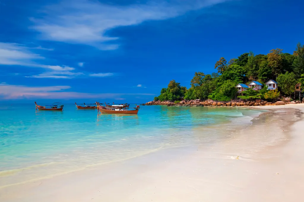 A breathtaking view of a pristine white-sand beach with turquoise water in Koh Lipe, Thailand. Several traditional longtail boats are anchored near the shore, and lush green hills with small bungalows line the beach, perfect for a luxury beach vacation.