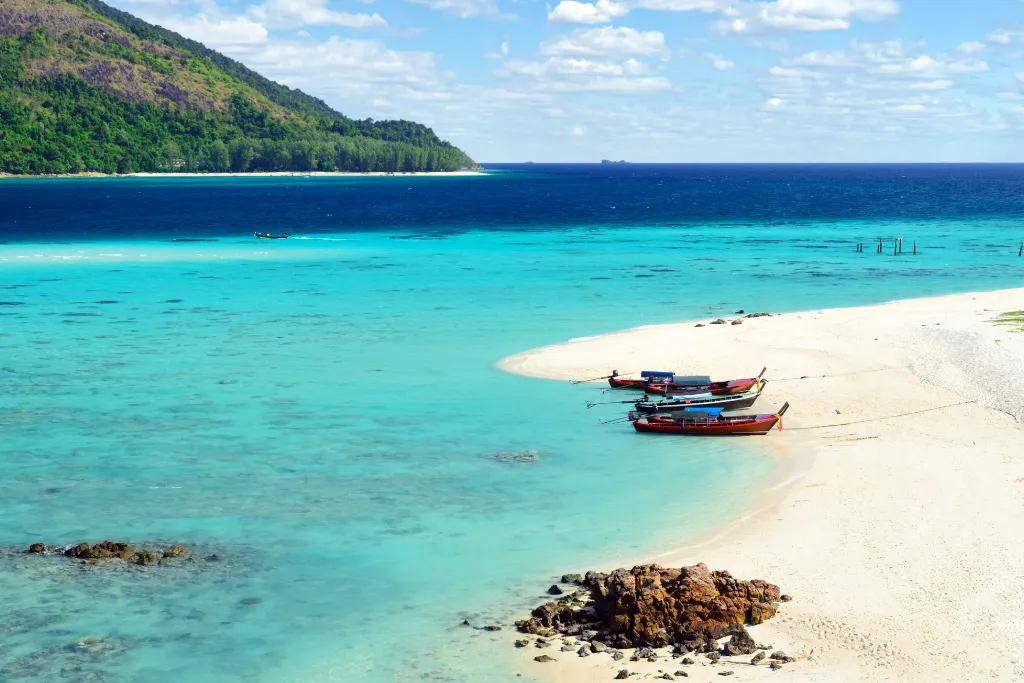An aerial-style view of a pristine white-sand beach with a unique curved shape on Koh Lipe, Thailand. The water transitions from a shallow, clear turquoise to a deep blue, with longtail boats resting on the shore. The image represents a luxury beach vacation and island-hopping.