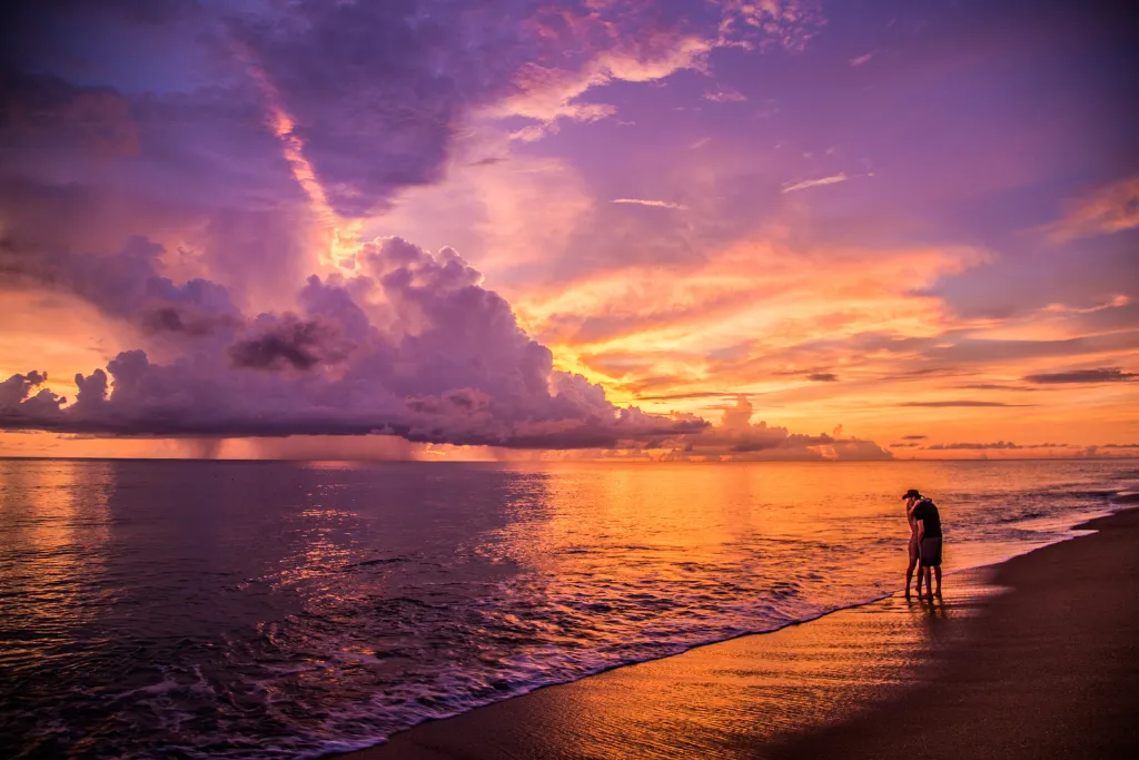 A romantic Khao Lak, Thailand, sunset with a couple standing on the beach, captured under a dramatic sky of vibrant purple and gold—the ultimate luxury travel experience.