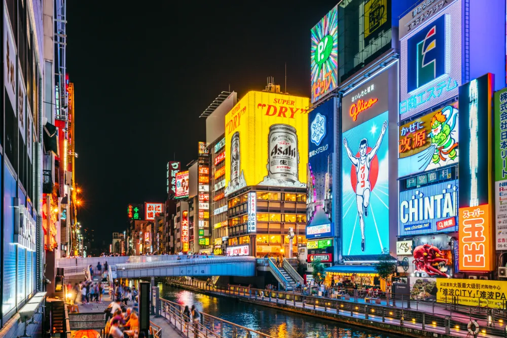 A dynamic night view of the Dōtonbori area in Osaka, featuring the famous Glico Running Man billboard and vibrant neon signs reflected in the canal. This scene is a perfect example of the exciting urban experiences available for luxury travel and MICE groups.