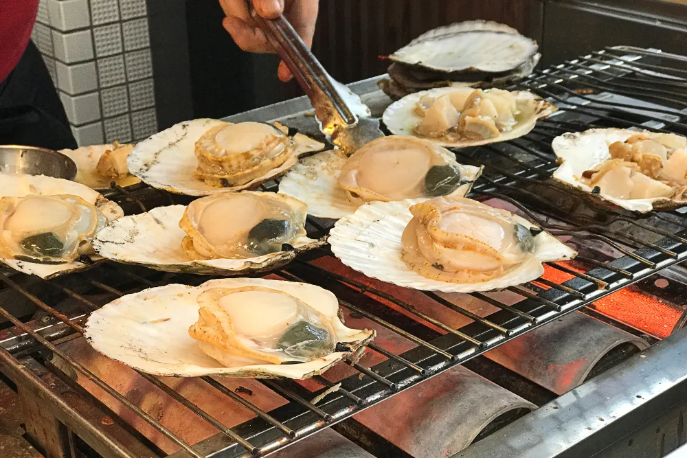  Fresh scallops are grilled at the famous Kuromon Ichiba Market in Osaka. This image captures the authentic culinary experience, perfect for a luxury food tour or a memorable MICE event.
