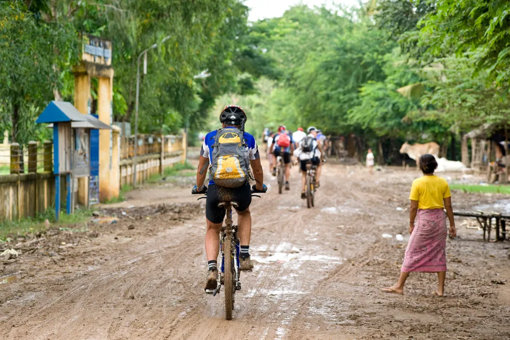 Tour group biking through a muddy, rural village in Cambodia, highlighting adventure travel and off-the-beaten-path experiences.