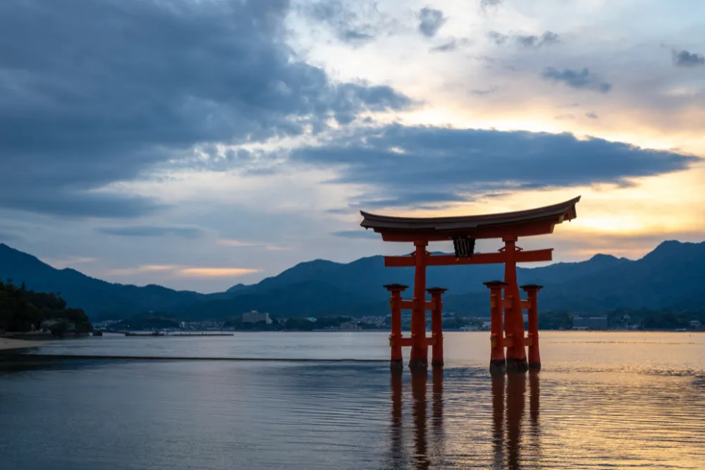 The iconic "floating" Itsukushima Shrine torii gate at high tide on Miyajima Island near Hiroshima, set against a dramatic sunset sky. This stunning UNESCO World Heritage site is a highlight of our exclusive MICE and corporate itineraries in Japan.