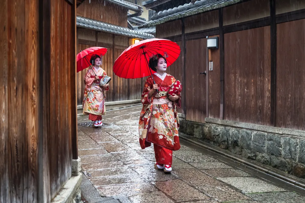 Two maiko or geisha with red umbrellas walking down a traditional street in Kyoto's Gion district. This image represents the authentic cultural experiences available for luxury tours in Japan.