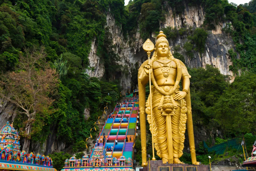 The massive gold statue of Lord Murugan stands beside the famous rainbow steps leading up to the Hindu temple within the limestone cave.