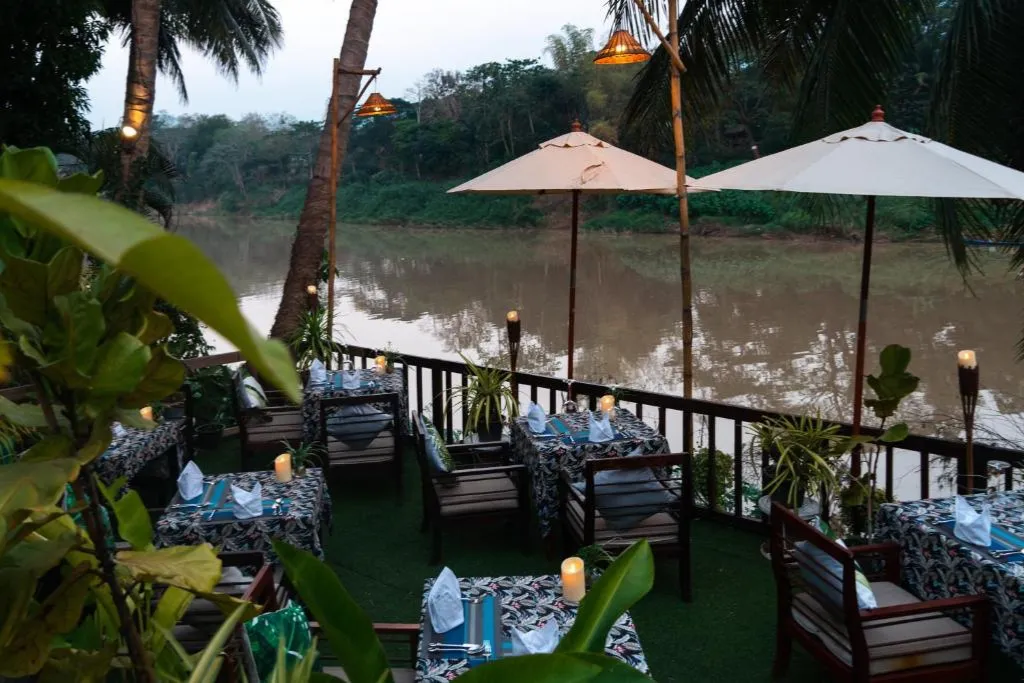 A romantic, candlelit riverside dining terrace in Laos overlooking the tranquil river at dusk. The perfect setting for a private luxury dinner on customized Laos tours and unforgettable Laos holidays.