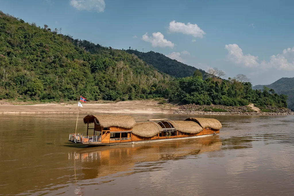 A luxury traditional slow boat with a thatched roof cruising the Mekong River, surrounded by lush green mountains. Perfect for high-end Laos river cruise experiences and exclusive Southeast Asia tours.