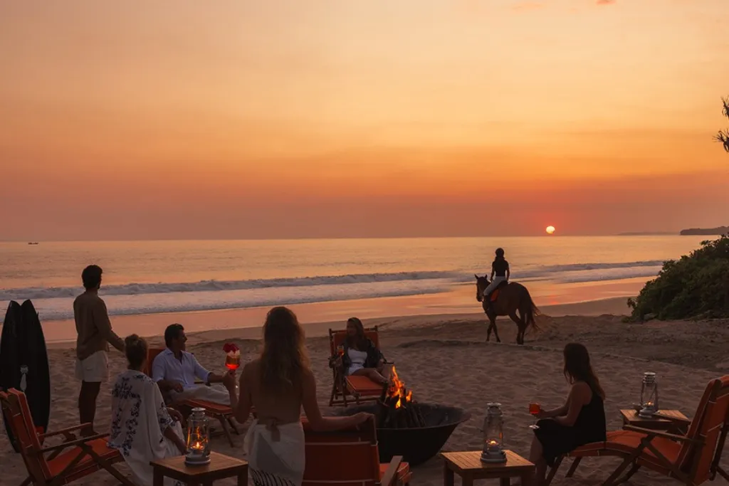 Idyllic scene of a stunning Sumba sunset over the ocean, with a group relaxing by a bonfire at the exclusive NIHI Sumba resort. Discover the height of luxury Indonesia holidays on Sumba Island.
