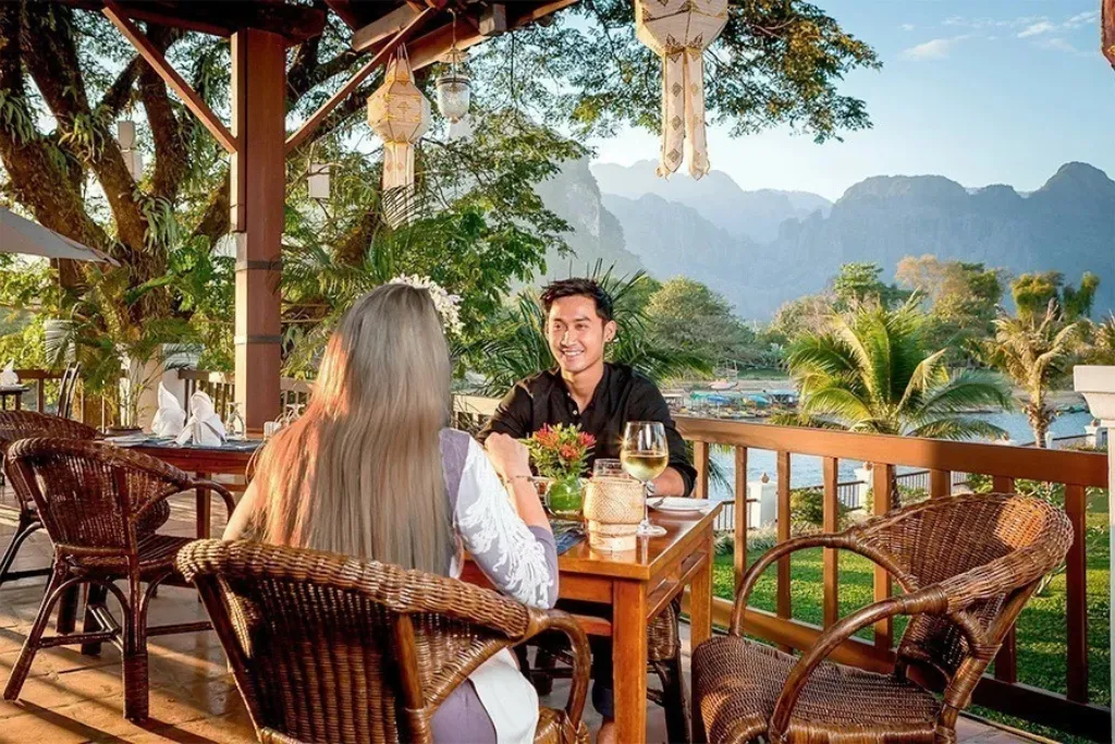 A couple enjoying a romantic, luxury dinner on a riverside terrace with a view of the beautiful karst mountains of Vang Vieng, Laos. Experience gourmet dining on your bespoke Laos tours and Laos holidays. Book your ultimate Laos travel experience with our expert Laos DMC.