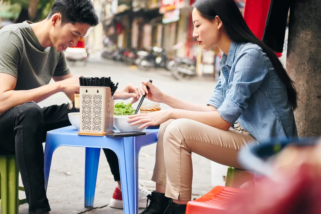 A couple enjoys authentic Vietnamese street food on plastic stools in Ho Chi Minh City (Saigon). Discover the best local flavors with our premium Vietnam food tours and culinary travel experiences during your luxury Vietnam holidays.
