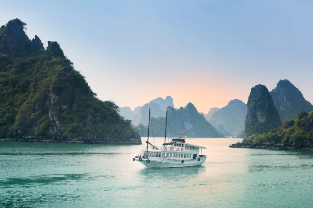 A luxury cruise boat sails on the emerald waters of Ha Long Bay, Vietnam, at sunset. Book unforgettable Vietnam tours and Vietnam holidays with our premium Vietnam DMC travel experts.