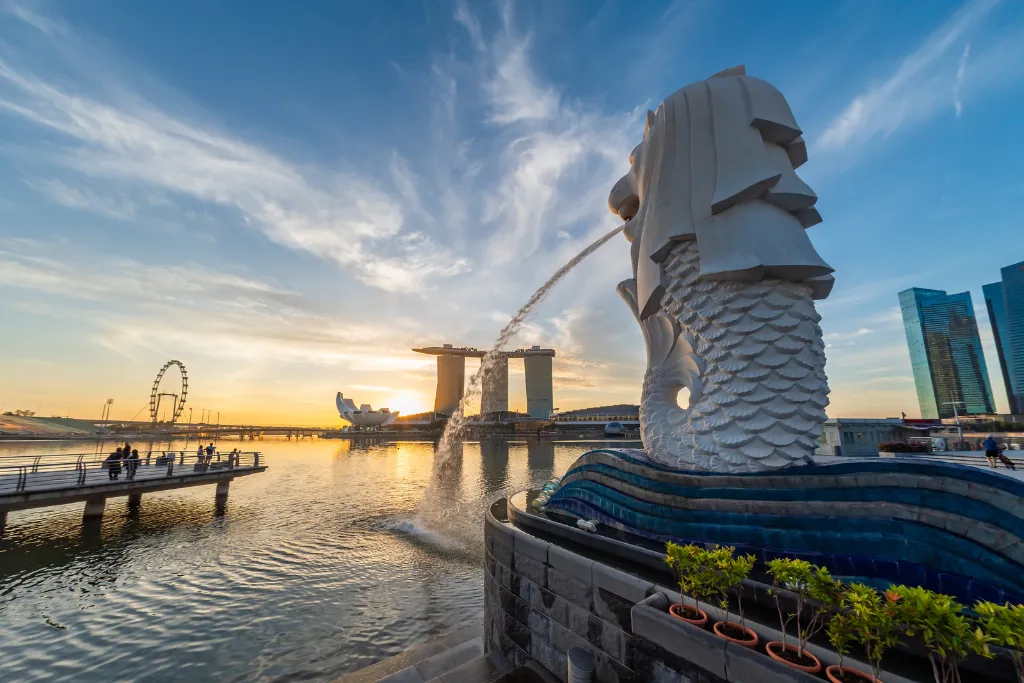 Iconic Merlion statue spouting water at sunset in Singapore, with Marina Bay Sands and the Flyer in the background.