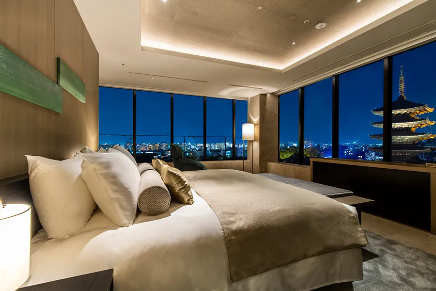 Luxury hotel suite in Kyoto, Japan, at night with a large, comfortable bed, minimalist decor, and panoramic window view of a traditional Japanese pagoda.