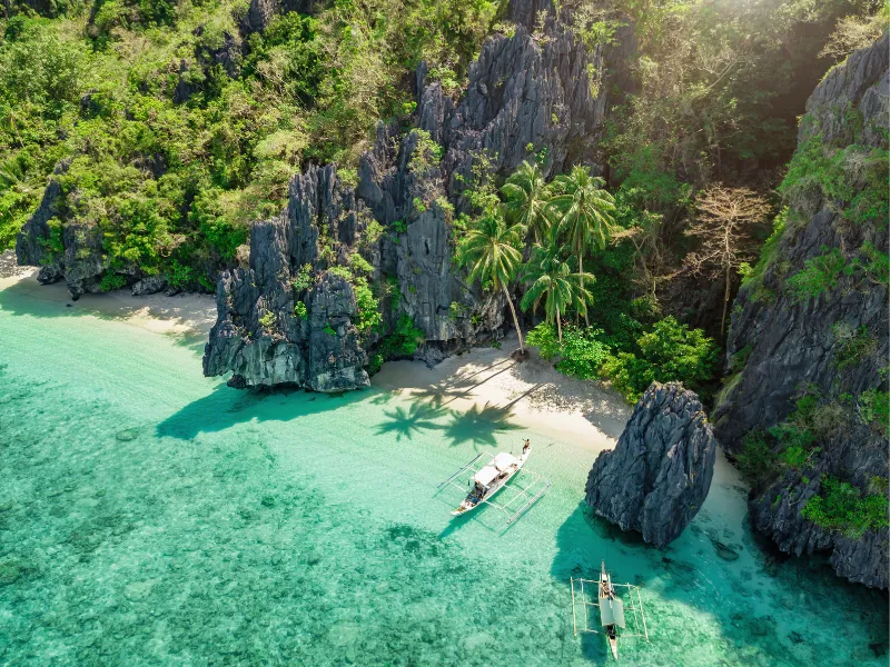An aerial view of a private, crystal-clear turquoise cove in Palawan, featuring traditional outrigger boats and dramatic limestone cliffs—curated by an expert Philippines travel operator for luxury island-hopping itineraries.