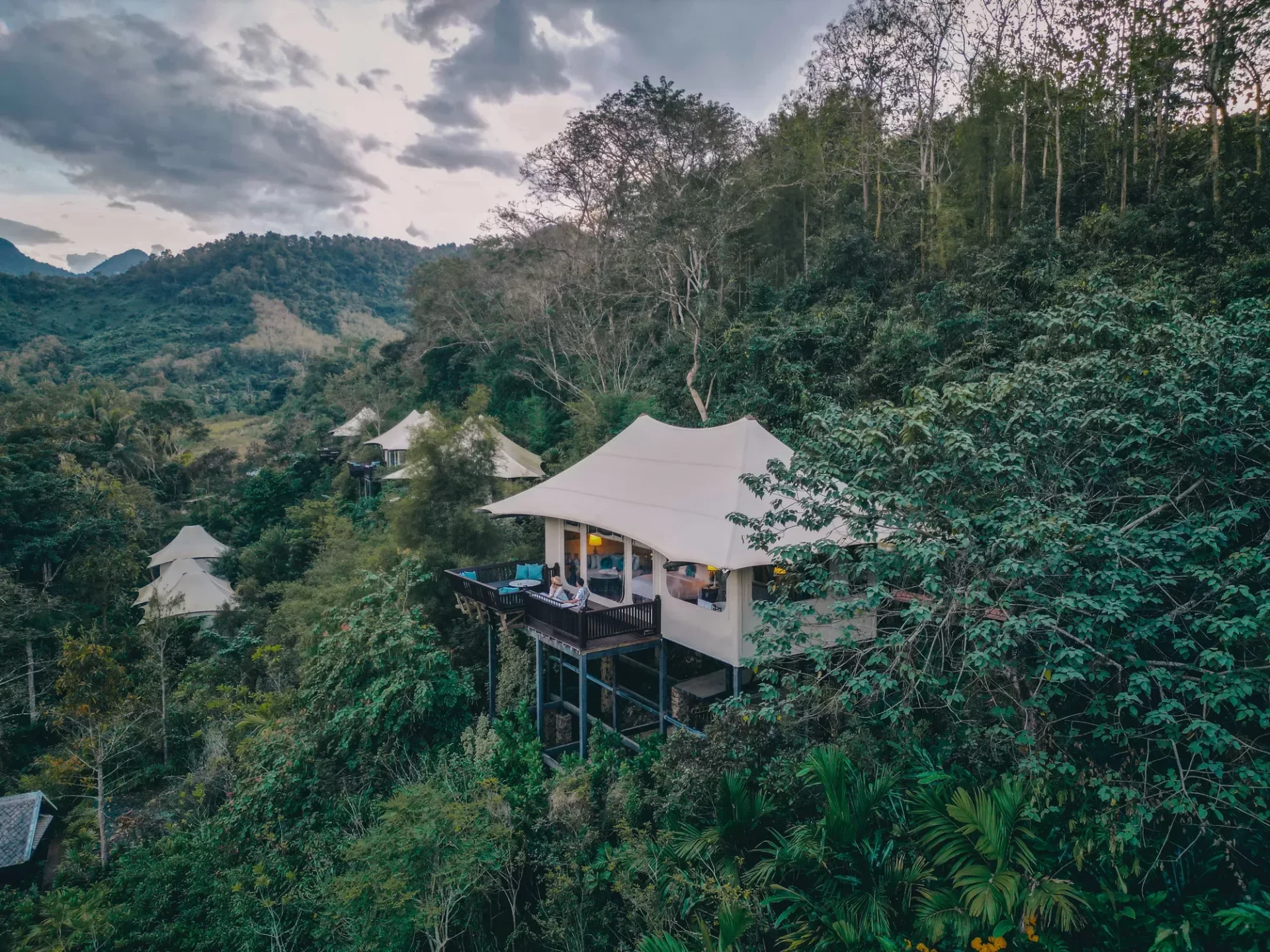 Aerial view of a luxury glamping tent perched on a jungle hilltop with a wraparound deck, surrounded by lush tropical forest and mountain scenery in Southeast Asia
