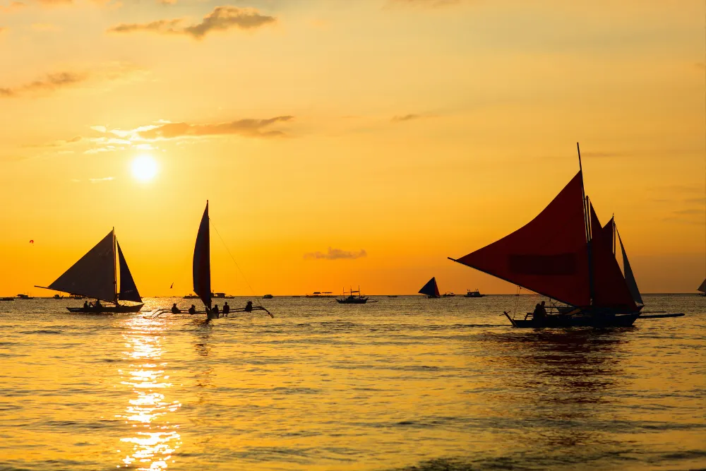 Silhouettes of traditional Filipino paraw outrigger sailboats with vibrant red sails gliding across golden sunset waters in Boracay, Philippines