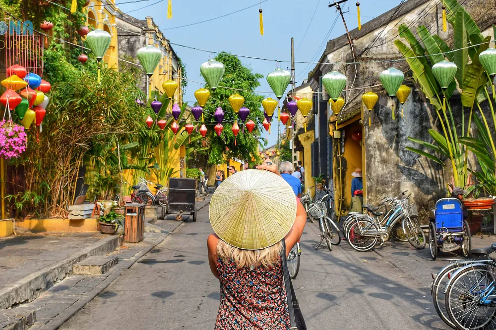 Traveller in a conical hat walking Hoi An's lantern-lined old town street, past yellow heritage buildings and bicycles