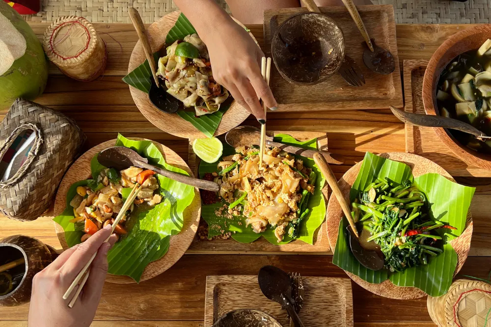 verhead view of a Lao meal served on banana leaves and woven trays, with sticky rice baskets and diners eating with chopsticks