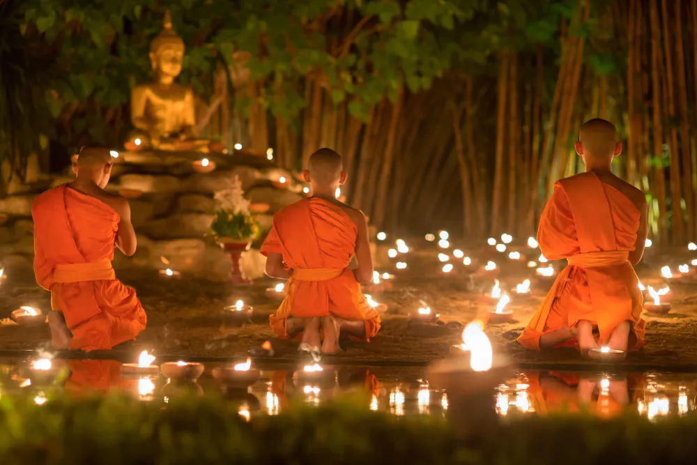 Young Buddhist monks kneeling in meditation amidst warm oil lamps during evening Thailand tours.