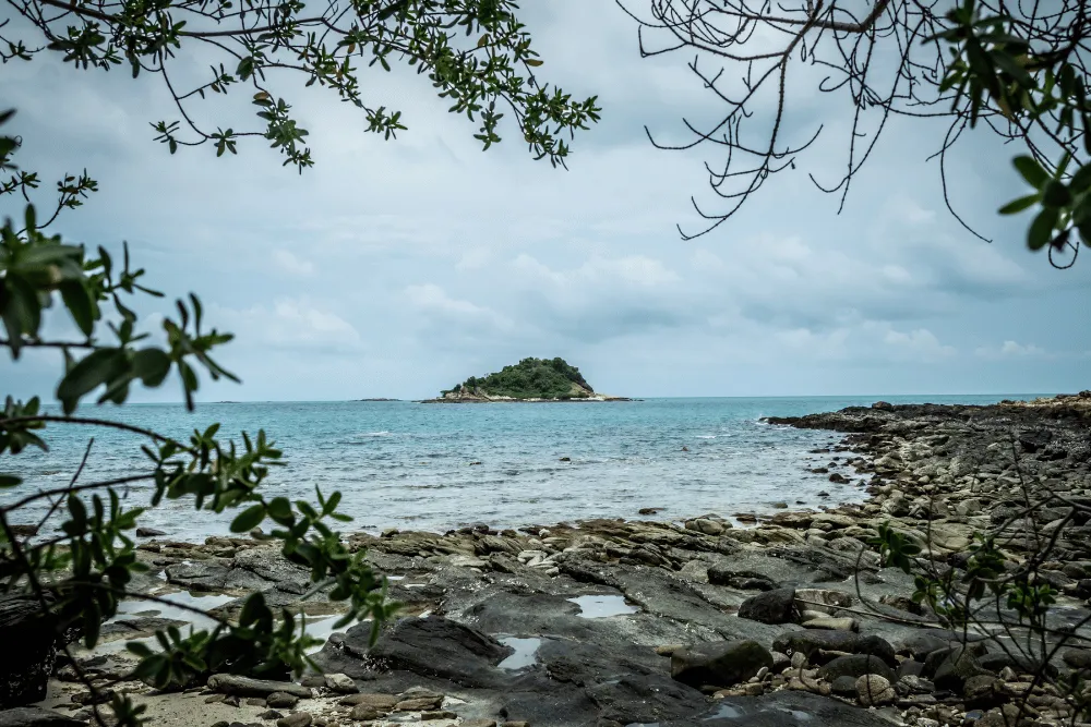 Scenic view of a small green islet across calm ocean waters from a rocky beach in Thailand.